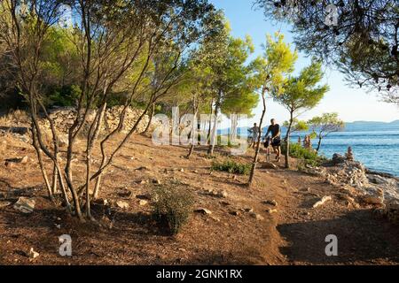 Podvrske, Murter, Croazia - 26 agosto 2021: Percorso tra pini presso la spiaggia rocciosa, e la gente che spinge le biciclette, nel tramonto estivo Foto Stock