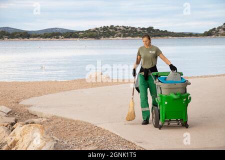 Vodice, Croazia - 28 agosto 2021: Giovane donna più pulita, che lavora per il servizio cittadino di Lec, al suo servizio di routine mattina pulizia della spiaggia Foto Stock