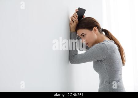 Triste depressione infelice attraente giovane donna caucasica con telefono si appoggia contro il muro bianco a casa, hanno problemi Foto Stock