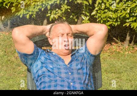 Ritratto di uomo maturo felice sotto spruzzi d'acqua in una giornata calda in giardino. Foto Stock