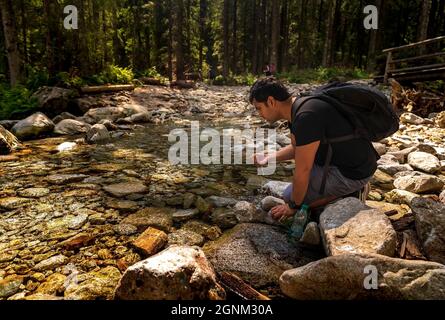 Uomo arabo indiano marrone con zaino seduto accanto al flusso d'acqua pulita o acqua potabile del fiume da esso, facendo una pausa durante il trekking o un'escursione nella foresta Foto Stock