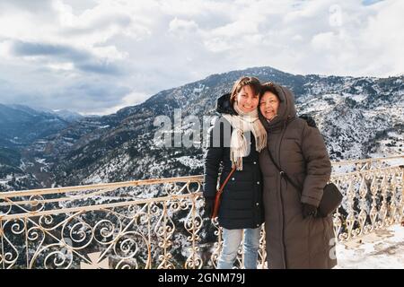 Ritratto invernale di madre e figlia sorridente e abbracciante con splendida vista sulle montagne innevate sullo sfondo, dalla terrazza del Monastero di Mega Spileon Foto Stock
