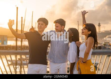 Giovani amici positivi e diversi abbracciando e ridendo mentre si prende selfie sul telefono cellulare in banchina durante le vacanze estive insieme al mare Foto Stock