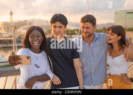 Gruppo di contenuti multirazziale giovani uomini e donne che guardano contenuti divertenti sul cellulare o prendere selfie mentre si levano in piedi insieme sul terrapieno in serata estiva Foto Stock