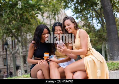Giovani amici femminili eleganti e diversificati con tazze di bevande da asporto che guardano le foto sul telefono cellulare mentre riposano dopo le visite turistiche durante le vacanze estive insieme in città Foto Stock