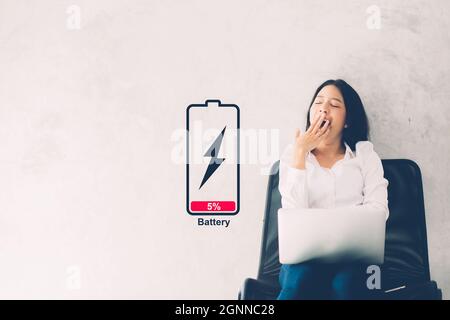 La giovane donna asiatica sbadigia al suo posto di lavoro con il computer portatile su sfondo cementizio, ragazza che lavora con sbadiglio e burnout con simbolo batteria bassa, stress Foto Stock