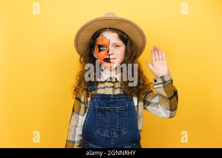 Ritratto di spaventoso divertimento vivo ragazza bambino con Halloween trucco, vestito in cappello e camicia, agitando la sua mano in gesto di ciao, isolato sul muro Foto Stock