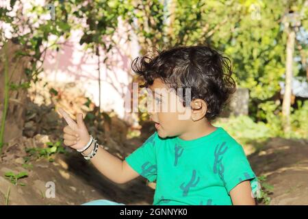 Primo piano di un piccolo bambino indiano che guarda un oggetto lontano con il gesto della mano e del dito. Piccolo ragazzo indù indiano che indossa verde Foto Stock