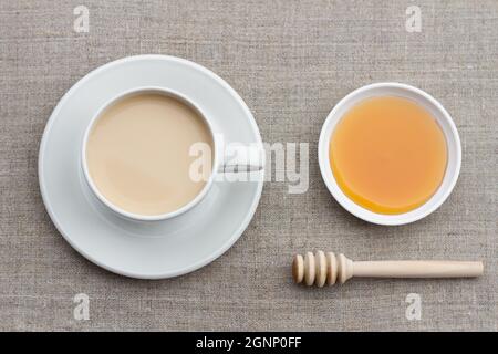 Tè piccante con latte in una tazza di porcellana bianca con cucchiaia di miele e miele su sfondo tessile Foto Stock