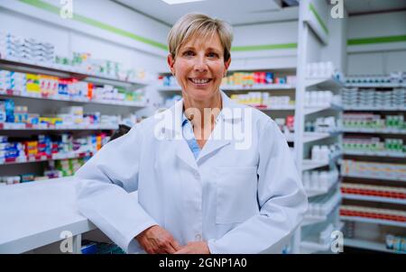 Sorridente farmacista caucasico senior appoggiandosi contro il banco in farmacia farmacia farmacia farmacia farmacia farmacia farmacia Foto Stock