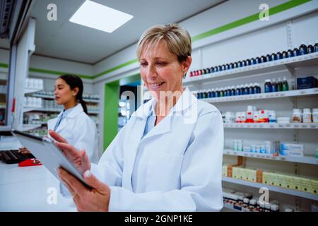 Il farmacista caucasico sorridente scorre attraverso la tavoletta digitale in piedi dietro il bancone in attesa di pazienti in farmacia Foto Stock