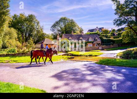 Due cavalli e cavalieri che attraversano ford attraverso il fiume tradizionale pietra costruito cottage villaggio Cotswold in background. Upper Slaughter Gloucestershire Regno Unito Foto Stock