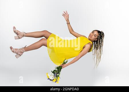 donna afroamericana in abito giallo levitating con bouquet isolato su bianco Foto Stock