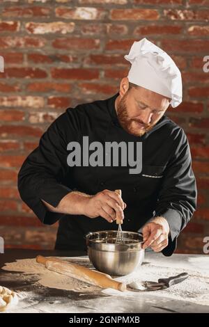 Ritratto di rosso-bearded uomo, chef, cuoco in nero uniforme cucina fa pasta di pane al caffè, cucina del ristorante. Concetto di dieta sana, nazionale Foto Stock