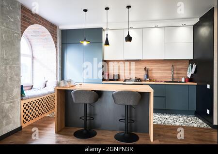 Vista della cucina contemporanea in bianco e grigio. Interrior di studio cucina con laminato in legno, grande finestra e molto lampade. Concetto di design elegante. Foto Stock
