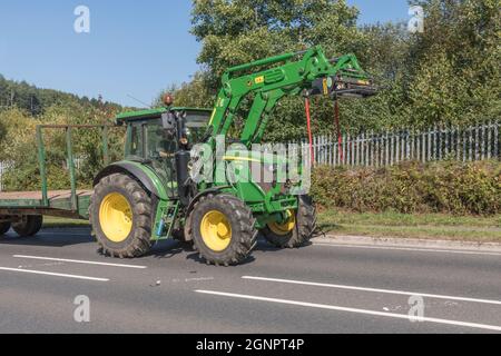 Trattore agricolo John Deere verde e familiare su strade rurali in salita aperte, con rimorchio agganciato. Il modello Deere con pneumatici grandi è 6115 RC. Foto Stock