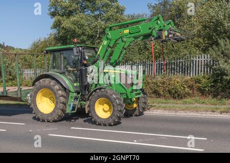 Trattore agricolo John Deere verde e familiare su strade rurali in salita aperte, con rimorchio agganciato. Il modello Deere con pneumatici grandi è 6115 RC. Foto Stock