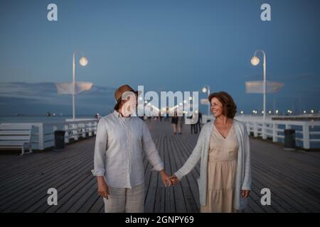Vista frontale della coppia anziana felice che cammina all'aperto sul molo dal mare al crepuscolo, tenendo le mani. Foto Stock