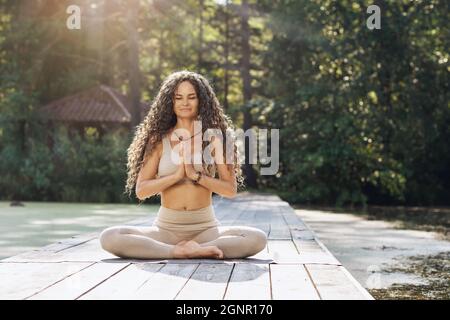 Una donna che pratica yoga, seduta su un tappeto in posizione di loto, medita su un ponte di legno vicino ad un laghetto, in una calda mattinata di sole Foto Stock