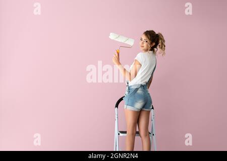 Bella donna è in piedi sulla scala e sta dipingendo il muro di colore rosa con un rullo sulla sua mano. Foto di alta qualità Foto Stock