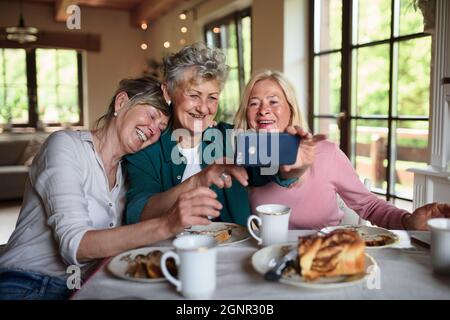 Amici anziani felici che hanno caffè e torta e prendere selfie in casa. Foto Stock