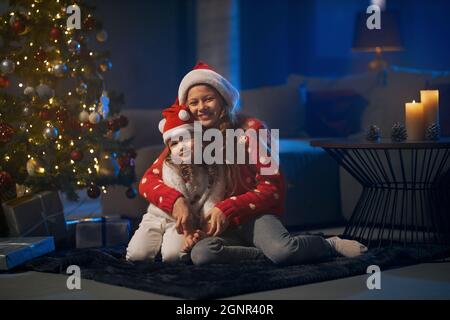 Sorelline felici a babbo natale cappelli seduti in abbracci sul pavimento e sorridenti con la macchina fotografica. Soggiorno scuro con candele brucianti e bellissimo albero di natale. Foto Stock