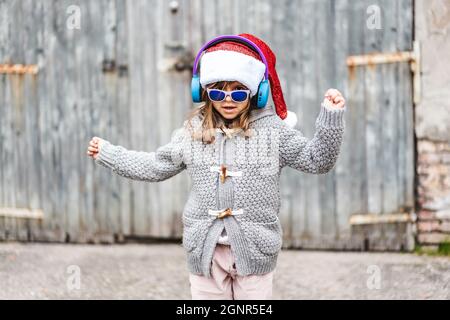 Divertente bambina che indossa cappello Babbo Natale e occhiali da sole che ballano contro uno sfondo di grunge in legno all'aperto nel periodo di Natale - buon Natale e Capodanno Foto Stock