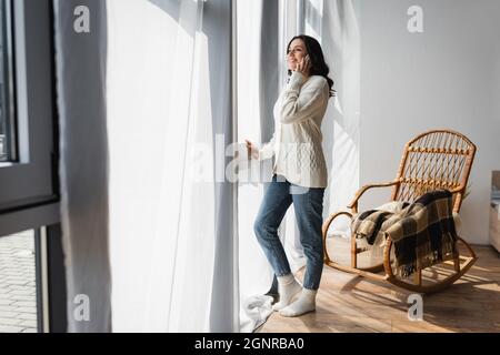 vista completa della donna in cardigan caldo in piedi vicino alla finestra e parlare sul cellulare Foto Stock