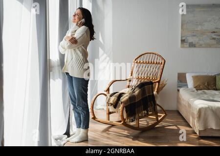 vista completa della donna in jeans e cardigan bianco che chiama su smartphone vicino alla finestra e poltrona in vimini Foto Stock