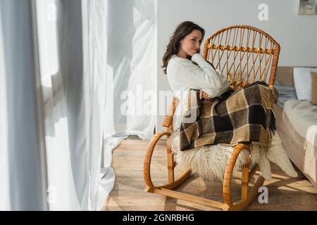 donna brunetta sorridente e guardando via mentre riposa in sedia a dondolo sotto coperta di plaid Foto Stock