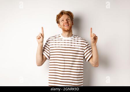 Buon giovane uomo con capelli rossi e la barba puntata, guardando in su con il sorriso sognante, controllando l'offerta speciale promo, sfondo bianco. Foto Stock