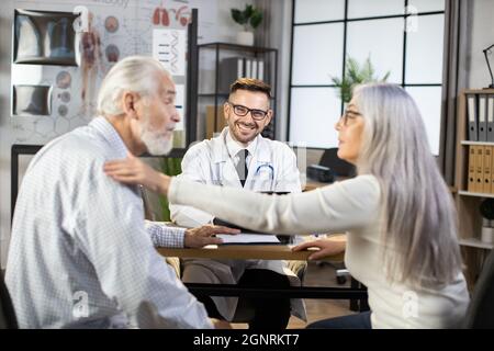 Medico maschile in occhiali e laboratorio bianco seduto alla scrivania e sorridente sulla macchina fotografica, fornendo consulenza medica per le coppie di età. Concetto di vecchia generazione e assistenza sanitaria. Foto Stock