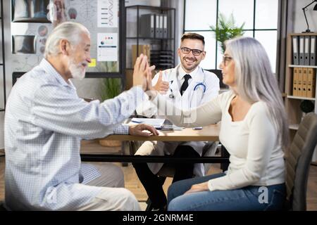 Coppia caucasica matura seduta con il medico maschile alla scrivania e dando alto cinque durante il controllo di successo. Bel medico sorridendo e mostrando il pollice fino alla fotocamera Foto Stock