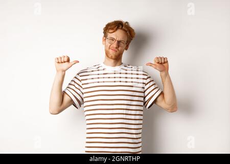 Giovane ragazzo sicuro in occhiali con capelli rossi, puntando a se stesso e guardando come professionale, sfondo bianco Foto Stock