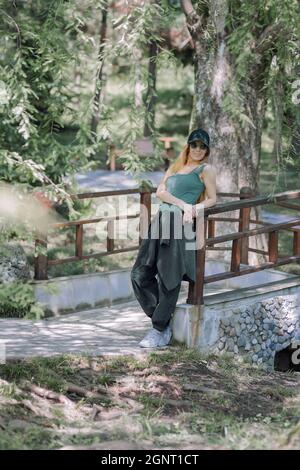 Sorridente ragazza sportiva in abito verde si erge vicino al ponte appoggiando il gomito sulla ringhiera nel parco estivo Foto Stock