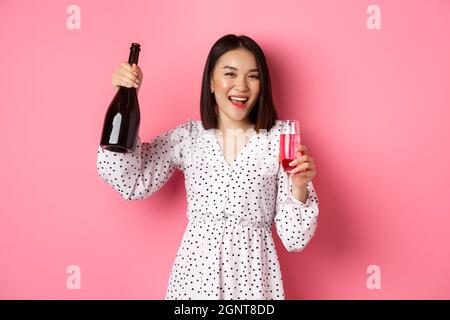 Felice donna asiatica che festeggia, divertendosi e festeggiando, versando un bicchiere di champagne e ridendo, in piedi su sfondo rosa Foto Stock