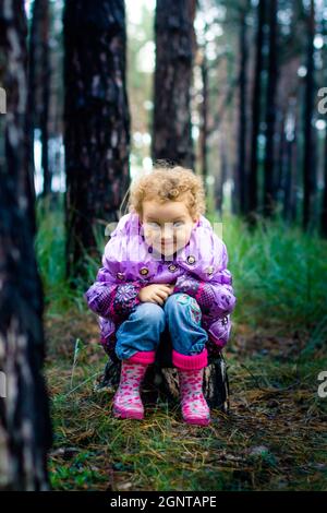 La bambina siede sul ceppo nei boschi. Ragazza curly in stivali di gomma. Il bambino sorrise slyly. Foto Stock