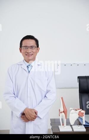 Ritratto verticale medio di medico asiatico maturo che indossa cappotto bianco e occhiali in piedi nel suo ufficio sorridendo alla macchina fotografica Foto Stock