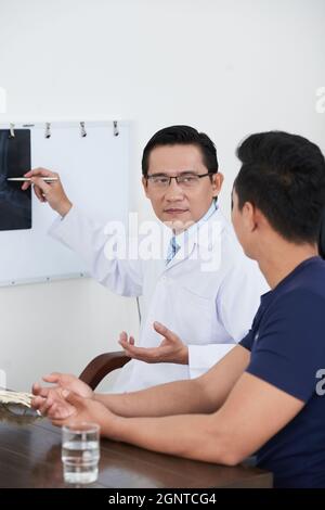 Ritratto verticale di medico asiatico maturo indossando cappotto bianco e occhiali seduti alla scrivania mostrando raggi X colpo al paziente spiegandogli qualcosa Foto Stock