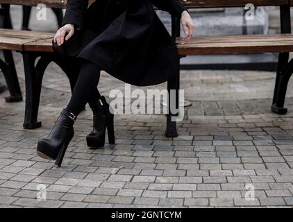 Donna che indossa cappotto nero e stivali in pelle e seduta su panca. Abbigliamento casual alla moda. Dettagli del look di tutti i giorni. Foto Stock