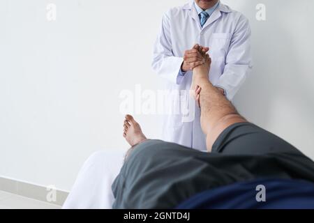 Shot orizzontale di medico osteopatico irriconoscibile che lavora con il paziente di sesso maschile che tiene la gamba facendo manipolazione dell'articolazione del piede Foto Stock