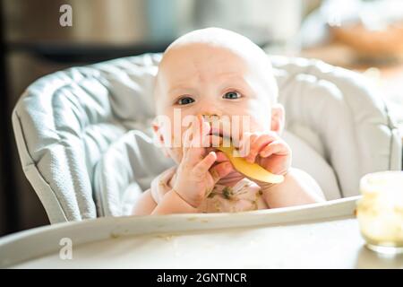 Bambina di 6 mesi a casa in seggiolone mangiare cibo gustoso a casa. Il bambino divertente con la puree di broccoli sani e sporchi tiene il cucchiaio nelle mani Foto Stock