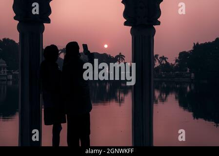 Silhouette di una coppia con volto nascosto scattare una foto selfie durante il tramonto vicino al lago di Pichola a Udaipur, Rajasthan Foto Stock