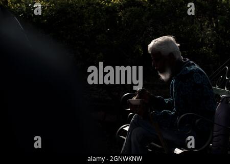 Foto di strada di un vecchio uomo con capelli bianchi e barba lunga con volto nascosto a causa della retroilluminazione mangiare il suo cibo mentre si siede su una panca Foto Stock