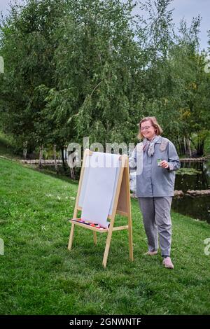 La donna felice si attinge al cavalletto con un pennello e vernici. Donna artista disegna la natura e gli alberi sulla carta vicino l'acqua sulla riva del fiume Foto Stock