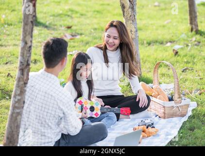 Felice asiatico giovane famiglia padre, madre e bambini che si divertono e si godono all'aperto insieme seduti sulla festa in erba con giocare Ukulele durante un Foto Stock