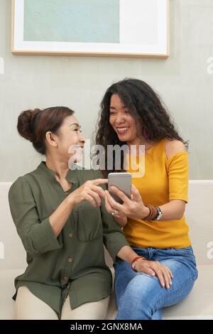 Felice giovane donna asiatica seduta sul divano e ridere con la madre alle foto sullo smartphone Foto Stock