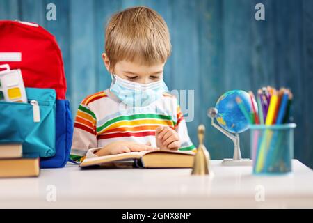 Ragazzo in maschera medica di protezione del viso in interni su sfondo blu. Bambino malato seduto al tavolo e leggere un libro. Pandemia di coronavirus. Foto Stock