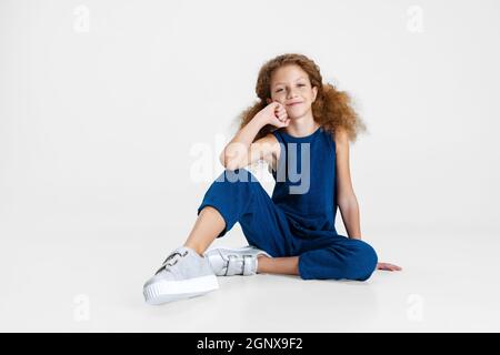 Ritratto di piccola ragazza carina caucasica in blu elegante tuta seduta sul pavimento isolato su sfondo bianco studio. Foto Stock