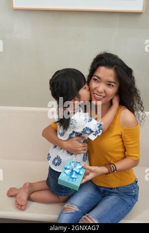 Ragazza asiatica carina con le trecce che abbraccia la madre intorno al collo e la bacia sulla guancia mentre si congratula per il giorno delle madri Foto Stock